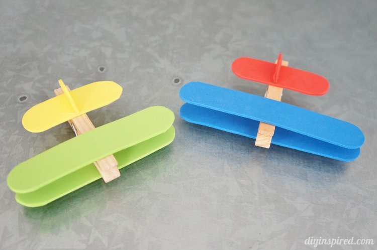 Two small, handmade toy airplanes made from colored popsicle sticks and clothespins on a gray textured surface. One plane is red and blue, and the other is yellow and green.