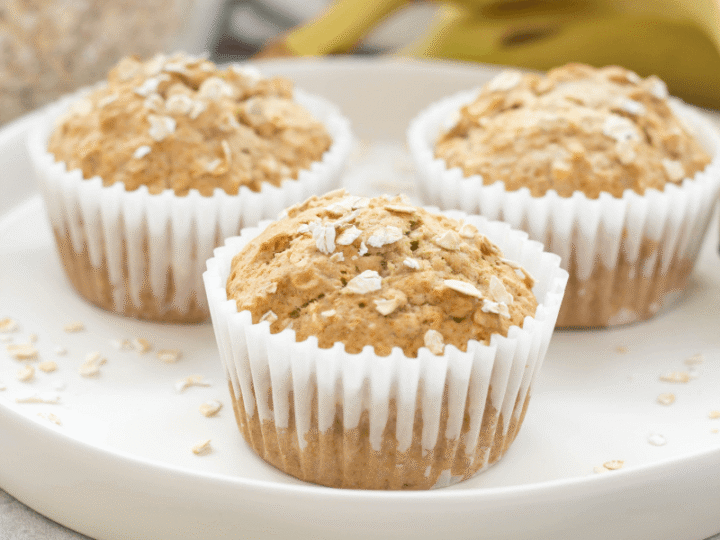 Three oatmeal muffins in white paper liners are displayed on a round, white plate. The muffins are topped with scattered oats, and there are a few oats on the plate. A banana is visible in the background.