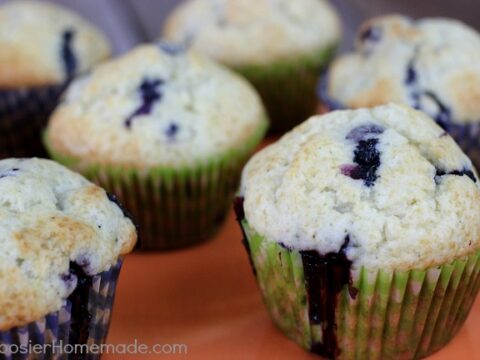 Close-up of five freshly baked blueberry muffins on an orange surface. The muffins are golden brown on top with visible blueberries and are wrapped in green and white muffin liners.