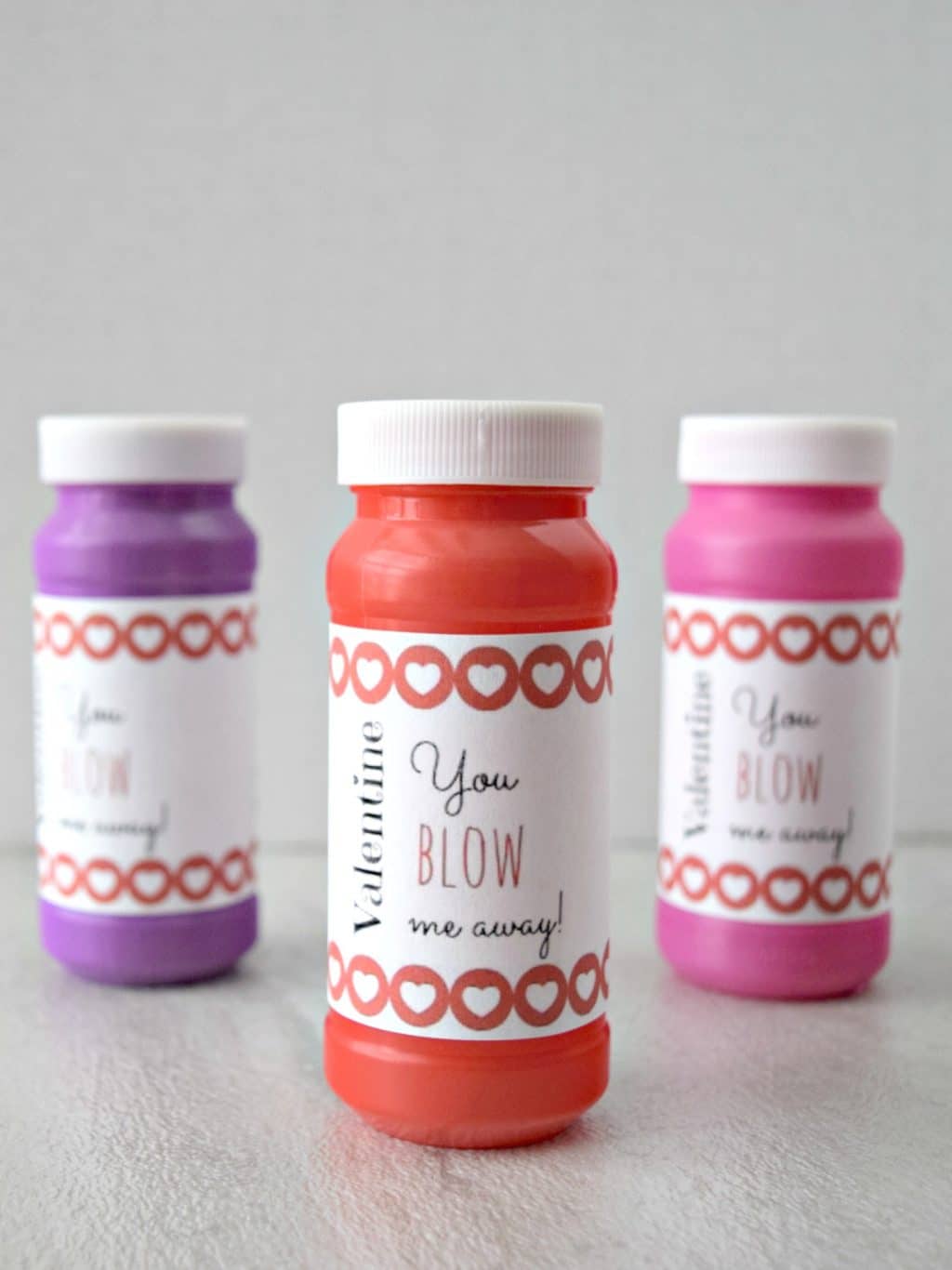 Three bottles of bubble solution are placed on a surface. Each has a label with heart patterns and the text Valentine You BLOW me away! The bottles are purple, red, and pink from left to right.
