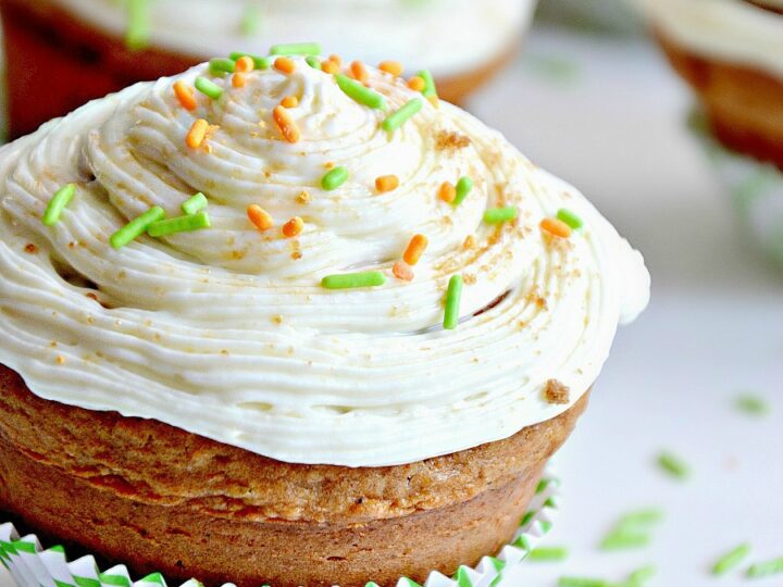 Close-up of a cupcake with swirled white frosting, topped with orange and green sprinkles. The cupcake is in a green and white striped liner. More cupcakes and sprinkles are blurred in the background.