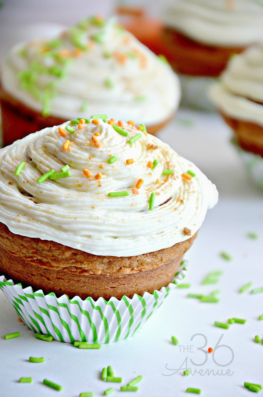 Close-up of a cupcake with swirled white frosting, topped with orange and green sprinkles. The cupcake is in a green and white striped liner. More cupcakes and sprinkles are blurred in the background.