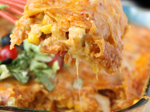 A close-up of cheesy enchilada casserole on a serving spoon with melted cheese, corn, and visible layers. In the background, the rest of the casserole and some garnished greens are visible in a glass dish. Text reads: Cheesy Enchilada Casserole - family fresh meals.