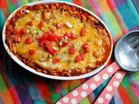 Oval dish of baked enchilada casserole topped with melted cheese, diced tomatoes, and green onions. It sits on a colorful checkered cloth next to a spoon and pink polka dot napkins.