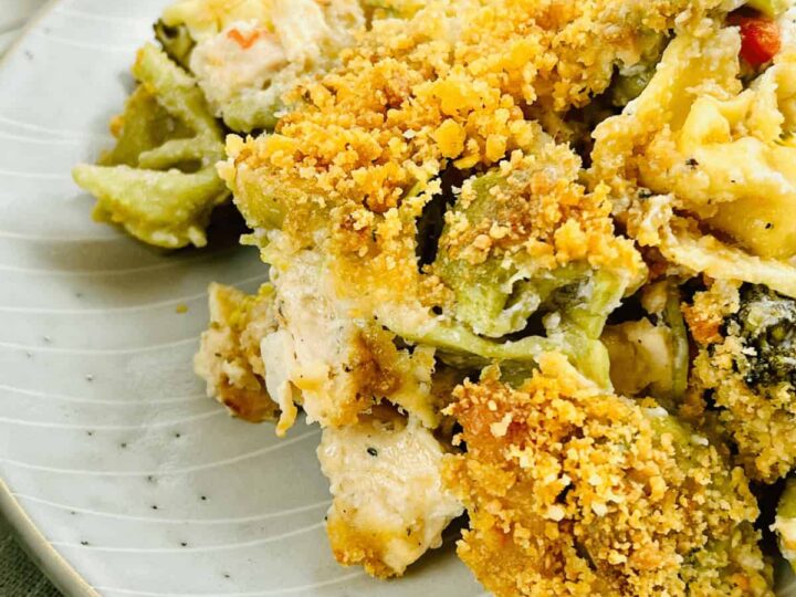 A plate of baked pasta with crispy breadcrumb topping. The pasta appears to be a mix of green and yellow varieties, showing a creamy sauce and bits of chicken or vegetables. The dish is served on a white speckled plate.