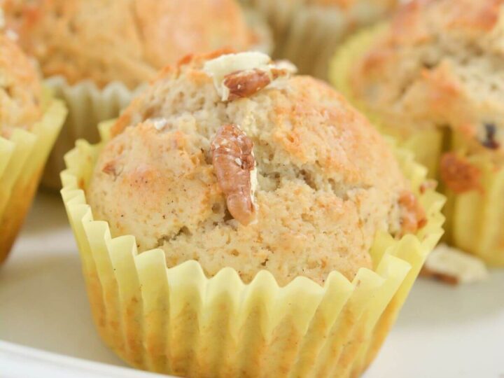 Close-up of a freshly baked muffin in a yellow paper cup, topped with chopped nuts. More muffins are visible in the background, resting on a white plate, showcasing a golden-brown texture.