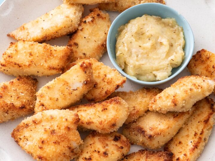 A plate of golden-brown, breaded chicken nuggets arranged around a small bowl filled with creamy dipping sauce. The nuggets appear crispy and are evenly spaced on the light-colored plate.