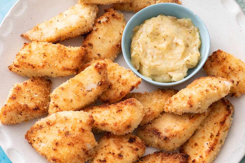 A plate of golden-brown, breaded chicken nuggets arranged around a small bowl filled with creamy dipping sauce. The nuggets appear crispy and are evenly spaced on the light-colored plate.