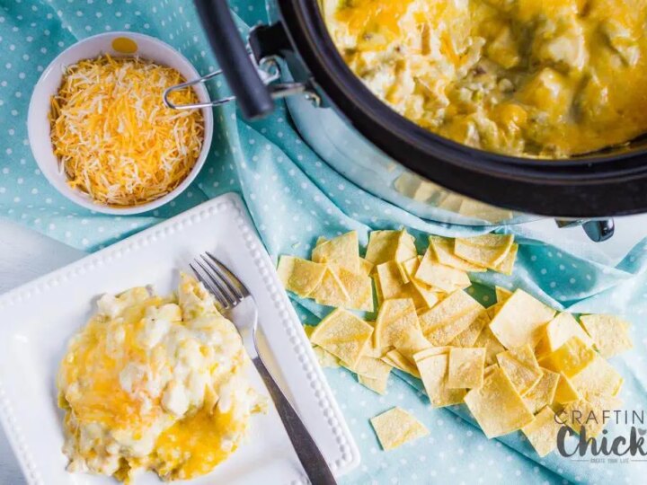 A table setting with cheesy potatoes in a crockpot on a blue polka dot cloth. A white plate holds a serving with a fork and knife beside it. Theres a bowl of shredded cheese and scattered tortilla chips on the side.