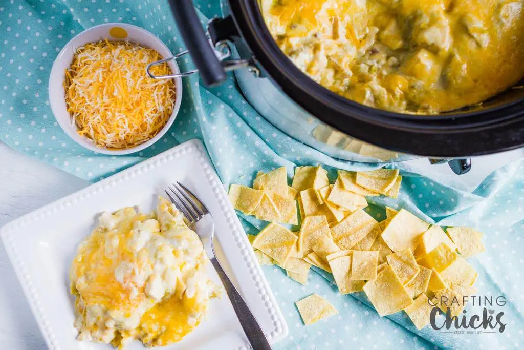 A table setting with cheesy potatoes in a crockpot on a blue polka dot cloth. A white plate holds a serving with a fork and knife beside it. Theres a bowl of shredded cheese and scattered tortilla chips on the side.
