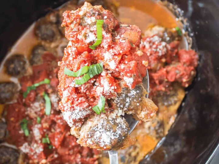A spoon holds meatballs topped with tomato sauce, grated cheese, and fresh basil over a slow cooker filled with more meatballs in sauce.