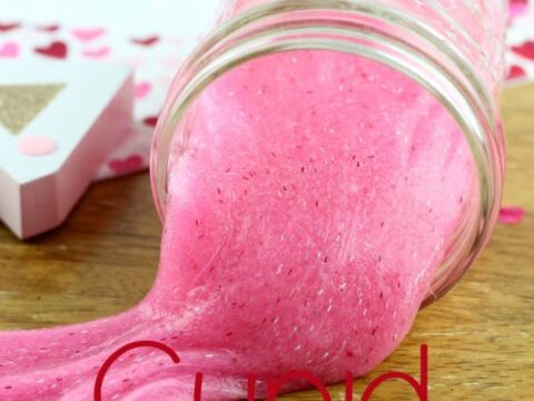 A glass jar tipped over on a wooden surface, spilling bright pink slime. The jar is labeled Cupid Snot (slime) and is surrounded by heart-themed decorations, including a small card with a heart design.