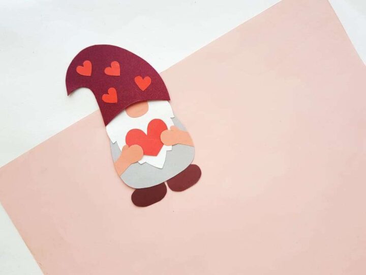A paper cutout of a gnome with a red hat decorated with red hearts. The gnome is holding a large red heart and is placed on a soft pink background, creating a whimsical and charming scene.