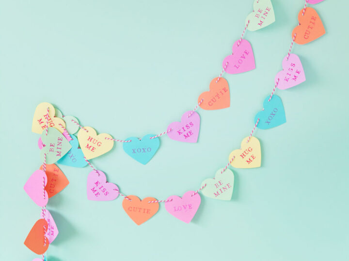 A colorful candy heart garland against a light green background. The hearts feature words like HUG ME, XOXO, and LOVE, creating a playful and festive decoration.