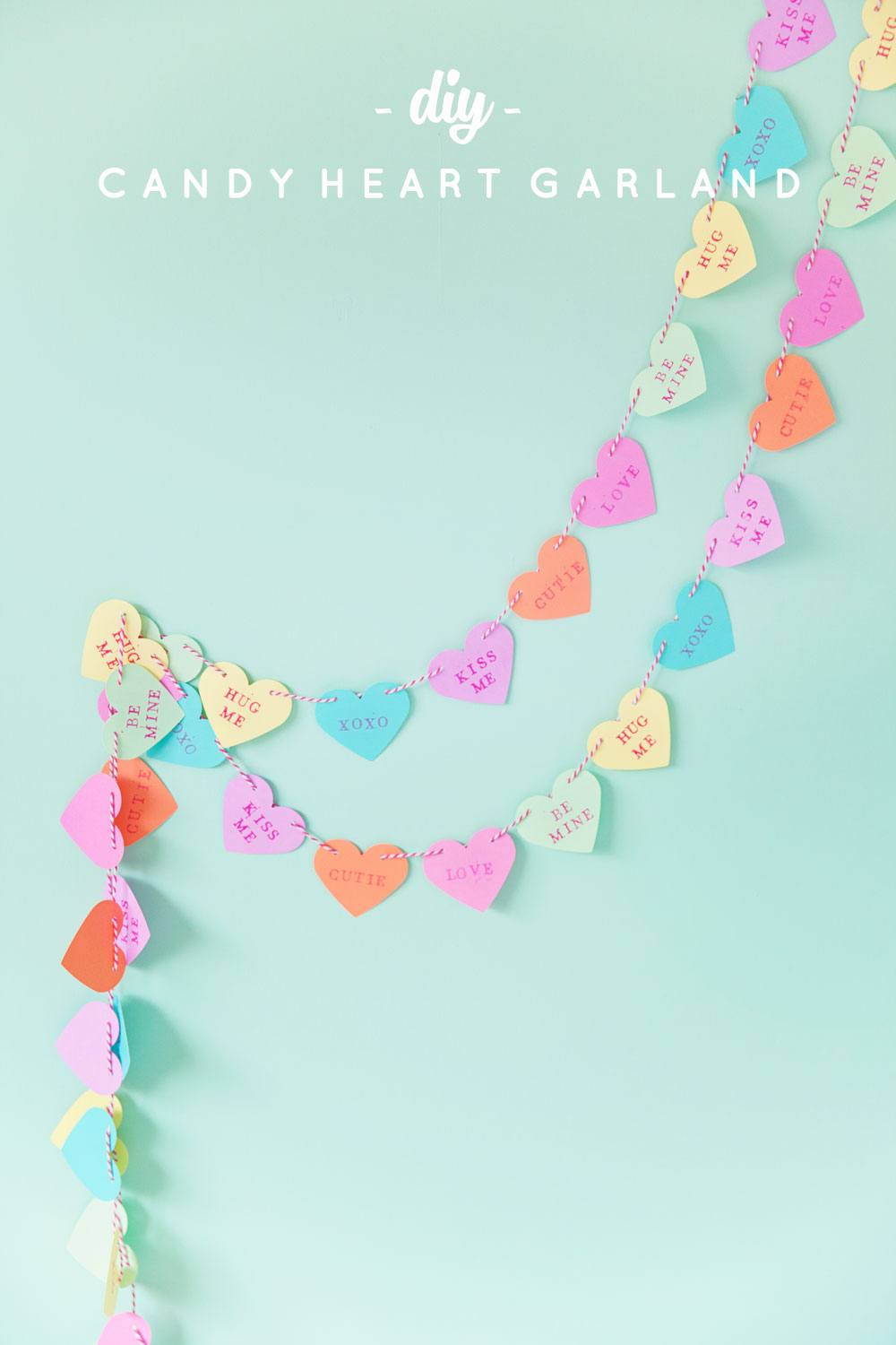 A colorful candy heart garland against a light green background. The hearts feature words like HUG ME, XOXO, and LOVE, creating a playful and festive decoration.