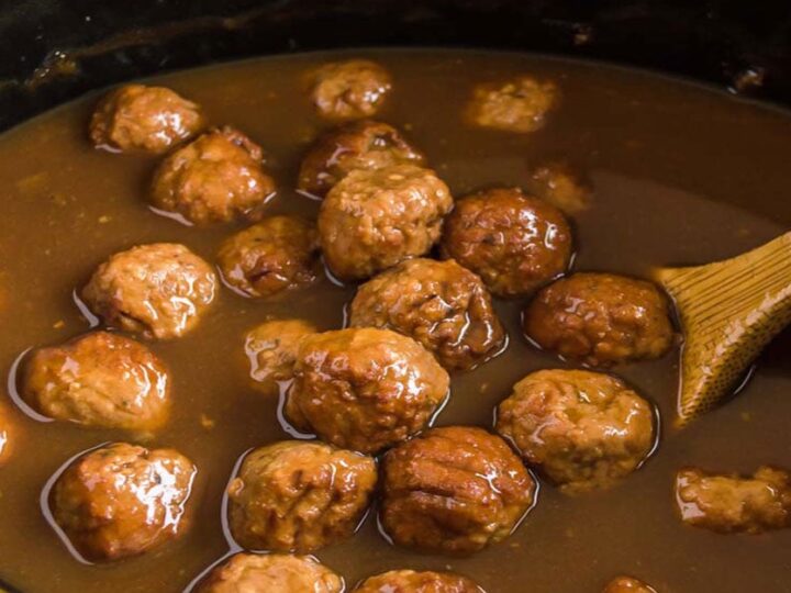 Meatballs simmering in a rich brown sauce inside a slow cooker. A wooden spoon is partially visible, used for stirring the dish. The scene suggests the meal is being prepared for serving.