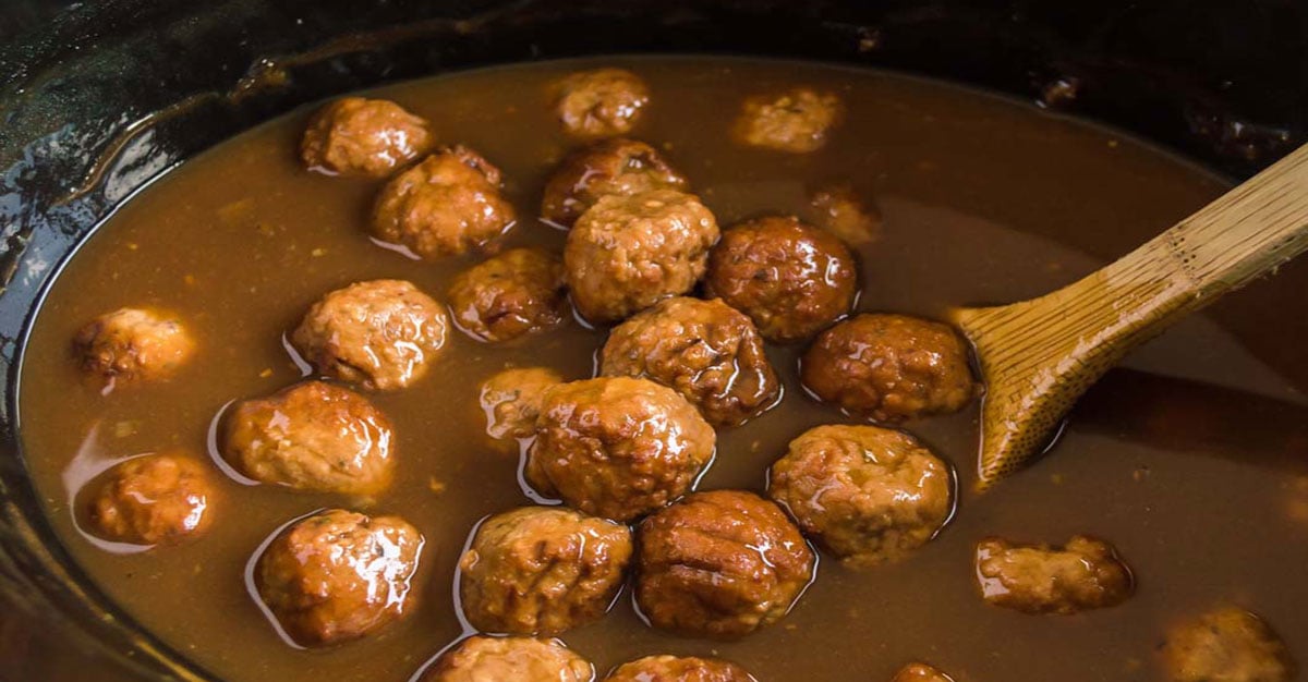 Meatballs simmering in a rich brown sauce inside a slow cooker. A wooden spoon is partially visible, used for stirring the dish. The scene suggests the meal is being prepared for serving.