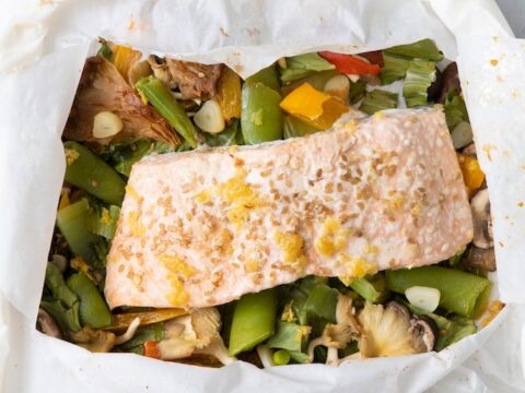 A piece of salmon fillet rests on a bed of mixed vegetables, including mushrooms, snap peas, and bell peppers, all wrapped in parchment paper. The presentation is fresh and colorful, ready to be baked or served.