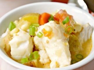 A white bowl filled with creamy fish chowder, containing chunks of fish, diced potatoes, carrots, and green onions as garnish. The bowl is on an orange and white checked surface, with a portion of a spoon visible in the background.