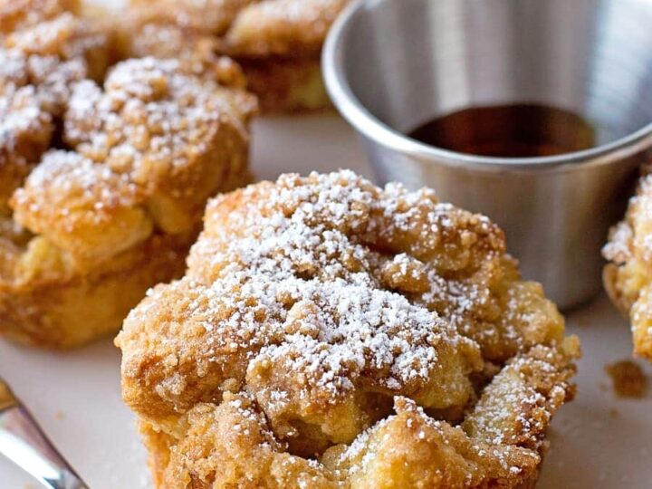 Muffin-shaped French toasts topped with powdered sugar sit on a white plate. A small metal cup of syrup and a fork are beside them. The plate is on a red and white striped cloth.