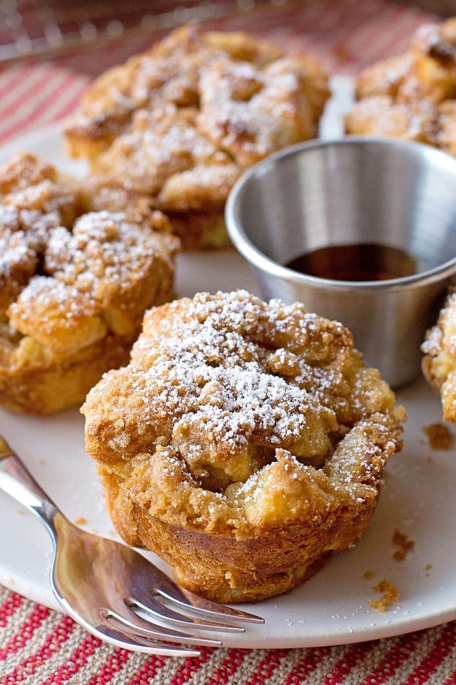 Muffin-shaped French toasts topped with powdered sugar sit on a white plate. A small metal cup of syrup and a fork are beside them. The plate is on a red and white striped cloth.