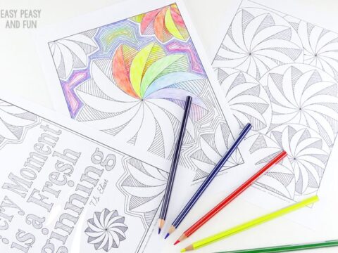 A flat lay of two patterned coloring pages, one partially colored with a rainbow design. Red, blue, green, and yellow colored pencils are scattered nearby. A sheet with a motivational quote is also visible.