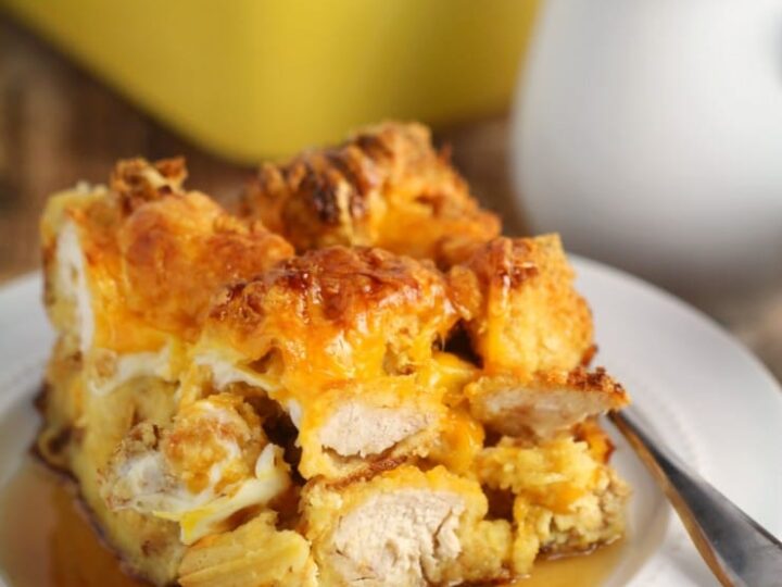 A slice of bread pudding topped with syrup is served on a white plate. A fork rests on a checkered napkin beside the plate. In the background, a yellow baking dish holds more of the dessert, next to a white pitcher.