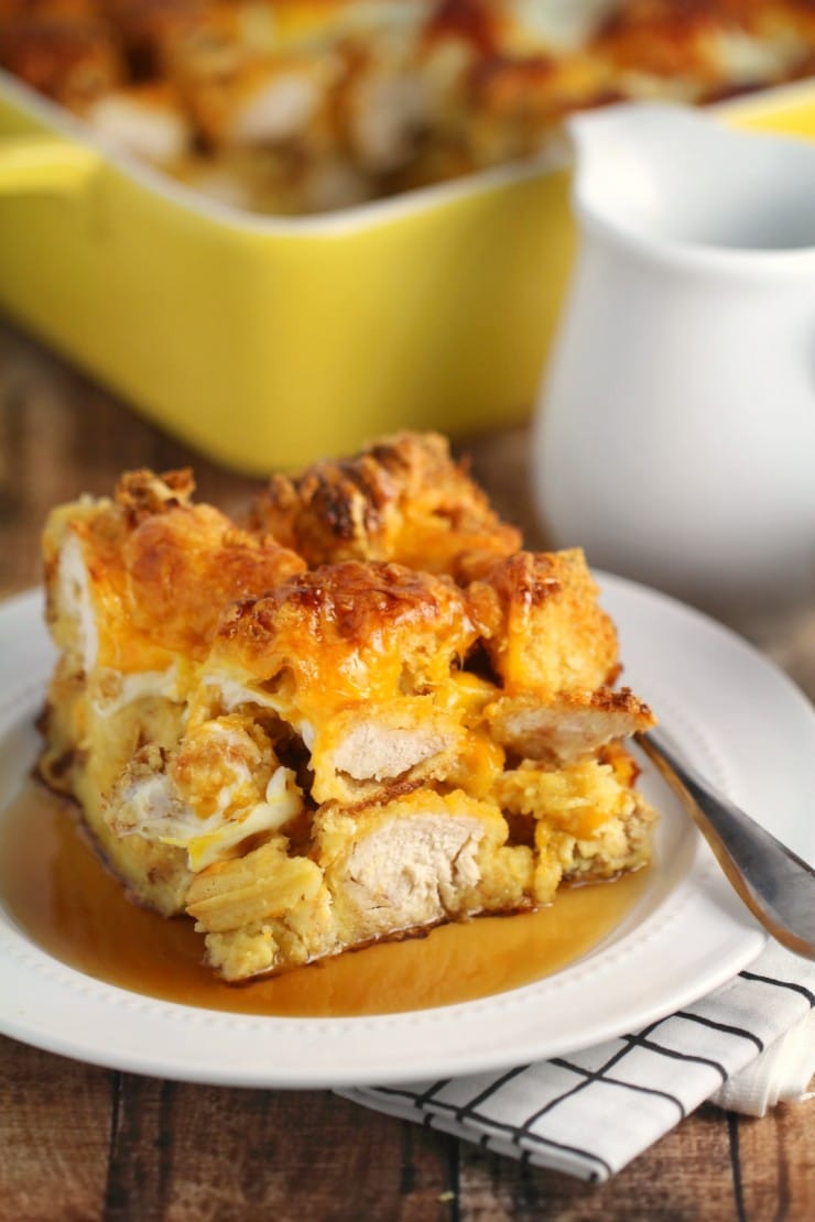 A slice of bread pudding topped with syrup is served on a white plate. A fork rests on a checkered napkin beside the plate. In the background, a yellow baking dish holds more of the dessert, next to a white pitcher.