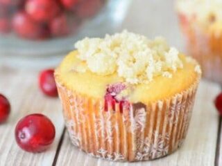 A cranberry muffin topped with crumbles sits on a wooden surface. Fresh cranberries are scattered around it. A glass bowl filled with more cranberries is slightly blurred in the background.