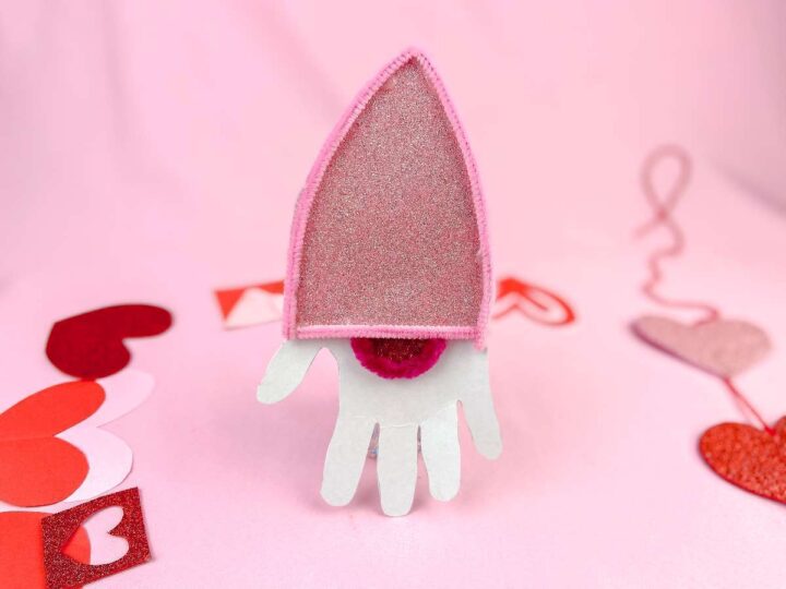 A pink felt hat with a glittery top sits on a backdrop decorated with paper hearts of various shades of pink and red. The design resembles a Bishops mitre and is surrounded by playful heart crafts.