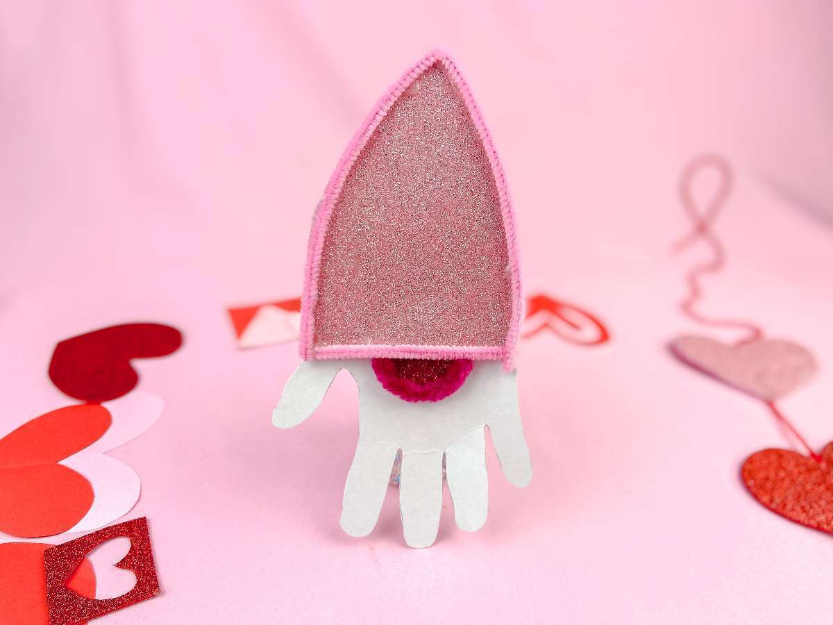 A pink felt hat with a glittery top sits on a backdrop decorated with paper hearts of various shades of pink and red. The design resembles a Bishops mitre and is surrounded by playful heart crafts.