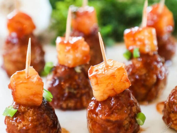 Meatballs topped with pineapple cubes are arranged on a plate. Each meatball is speared with a toothpick, and garnished with small green onion pieces. The background features assorted greens.