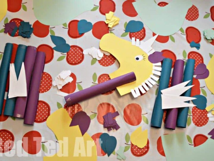 Colorful paper craft materials are arranged on a table with an apple-patterned tablecloth. The materials include paper shapes resembling a horses head with a mane, rolled paper tubes, and various flowers and leaves in vibrant colors.