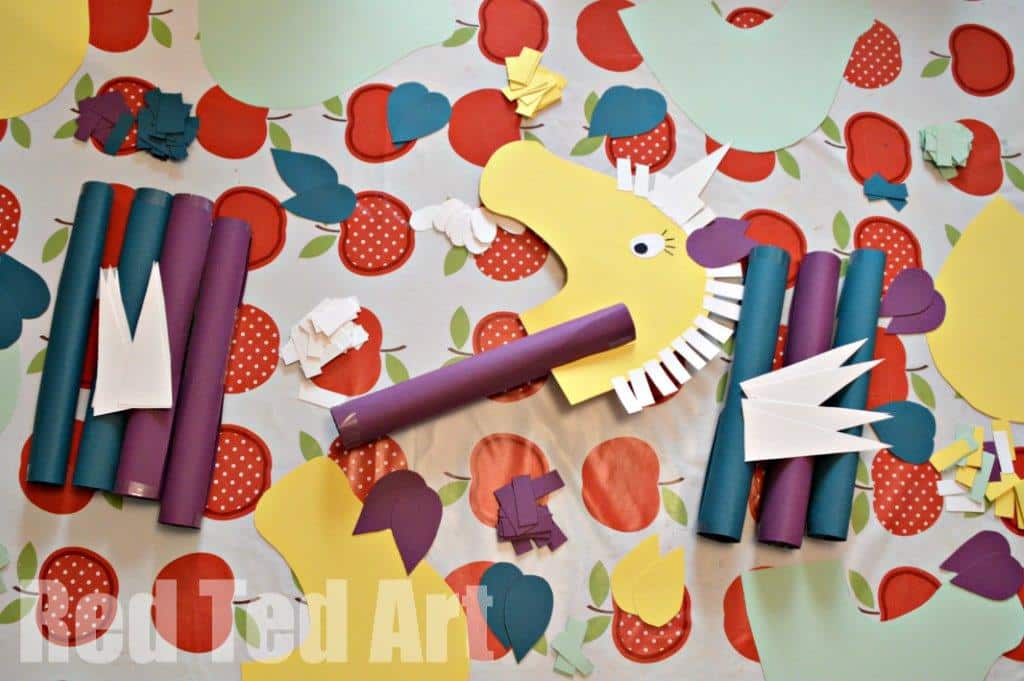Colorful paper craft materials are arranged on a table with an apple-patterned tablecloth. The materials include paper shapes resembling a horses head with a mane, rolled paper tubes, and various flowers and leaves in vibrant colors.