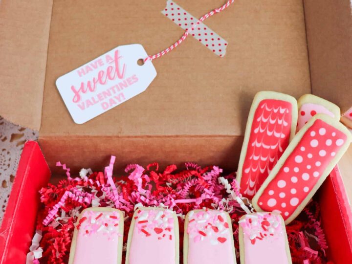Open cardboard box with decorated cookies. Five rectangular cookies spell LOVE in pink icing, surrounded by red and pink decorative paper. A tag reads Have a sweet Valentines Day.