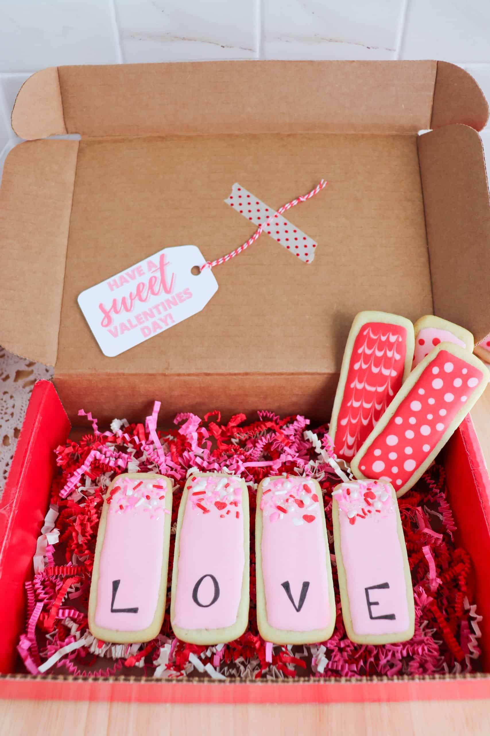 Open cardboard box with decorated cookies. Five rectangular cookies spell LOVE in pink icing, surrounded by red and pink decorative paper. A tag reads Have a sweet Valentines Day.