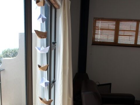 A series of paper boats hanging in a line next to a window with light curtains. The room has a black sofa, a small stove, and blinds covering the window, with some greenery visible outside.