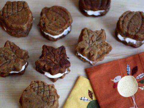 Autumn-themed cookies shaped like leaves and pumpkins, filled with a creamy white filling, are arranged on a light wooden surface. Two colorful napkins with embroidered designs are placed nearby.