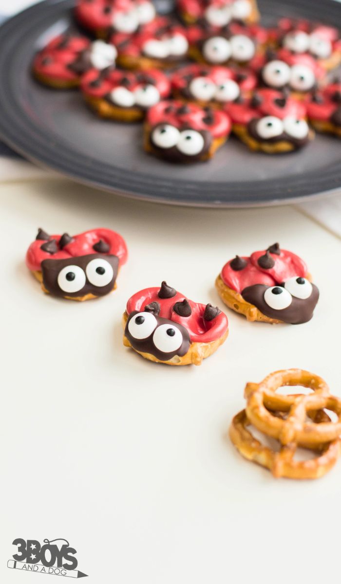 A plate of pretzels decorated to resemble charming lovebugs with red and brown icing and candy eyes. Three are in focus on a white surface, accompanied by two plain pretzels.