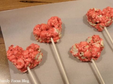 Four pink heart-shaped popcorn treats on sticks are laid out on a parchment sheet. The treats have a glossy coating and are placed in a neat arrangement.