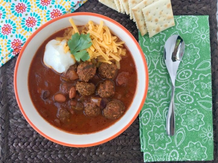 A bowl of chili with meatballs and kidney beans, topped with sour cream, shredded cheese, and cilantro. Its placed on a woven mat. A green patterned napkin, spoons, and crackers are on the side. A colorful cloth is partially visible.