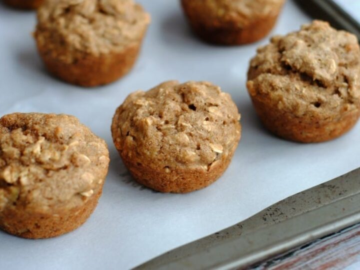 Close-up of several freshly baked oat muffins on a baking sheet lined with parchment paper. The muffins have a golden-brown, textured surface, indicating a hearty, rustic appearance.