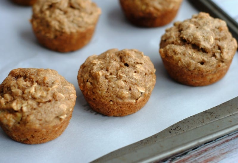 Close-up of several freshly baked oat muffins on a baking sheet lined with parchment paper. The muffins have a golden-brown, textured surface, indicating a hearty, rustic appearance.