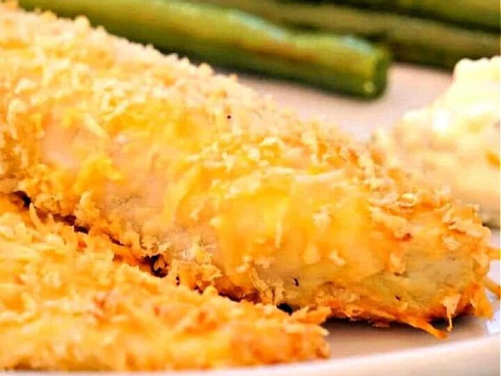 Close-up of a plate featuring breaded and baked fish fillets with a golden, crispy coating. In the background, there are green beans and a side of creamy sauce.