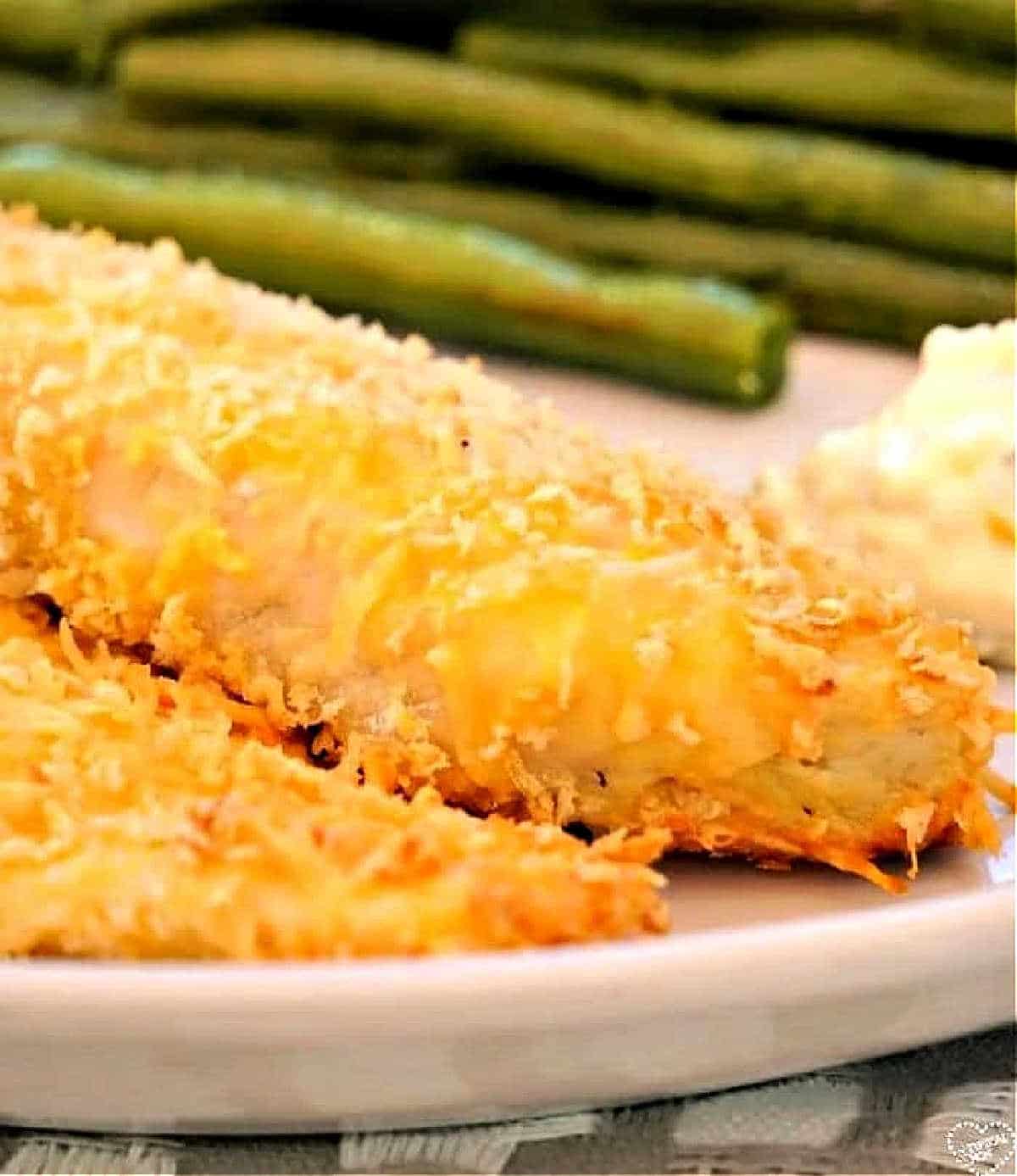 Close-up of a plate featuring breaded and baked fish fillets with a golden, crispy coating. In the background, there are green beans and a side of creamy sauce.