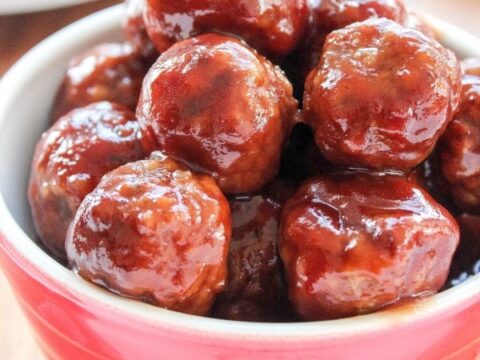 A red bowl filled with glazed meatballs, placed on a wooden surface. The meatballs are coated in a glossy brown sauce, appearing appetizing and well-cooked.