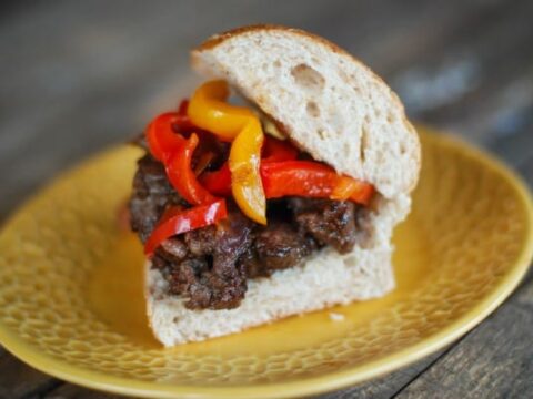 Sandwich on a yellow plate with sliced beef and colorful bell peppers, red and yellow, on a wooden table.
