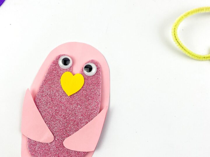 A pink and purple bird made of paper with googly eyes, a yellow heart-shaped beak, and red feet on a white surface. Colorful paper and a yellow pipe cleaner are partially visible in the corners.