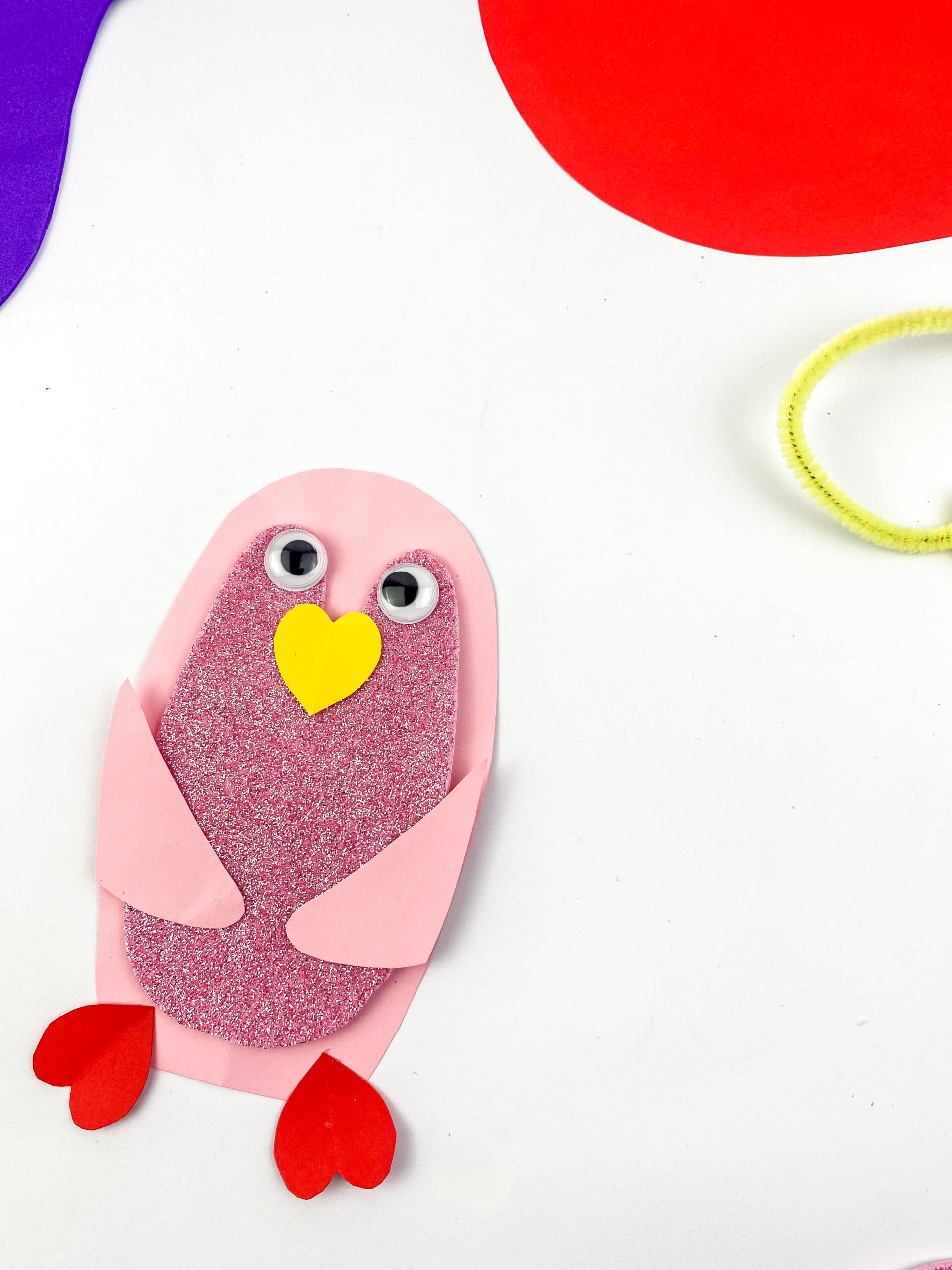 A pink and purple bird made of paper with googly eyes, a yellow heart-shaped beak, and red feet on a white surface. Colorful paper and a yellow pipe cleaner are partially visible in the corners.