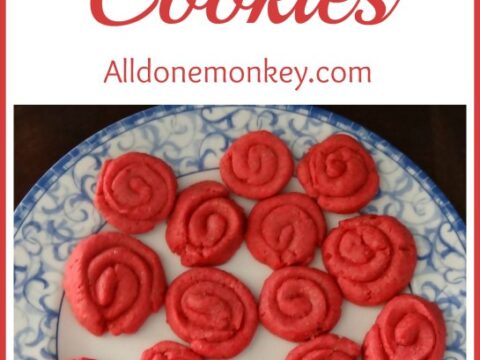 A decorative plate holds rose-shaped cookies in vibrant red. The white rim features a blue floral pattern. Header text reads Rose Cookies, and a website name is listed below.